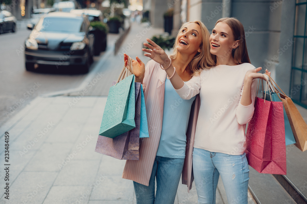 Look over here. Portrait of beautiful senior woman and young lady holding shopping bags while standing on street. Smiling girl pointing at something Look over here. Portrait of beautiful senior woman and young lady holding shopping bags while standing on street. Smiling girl pointing at something