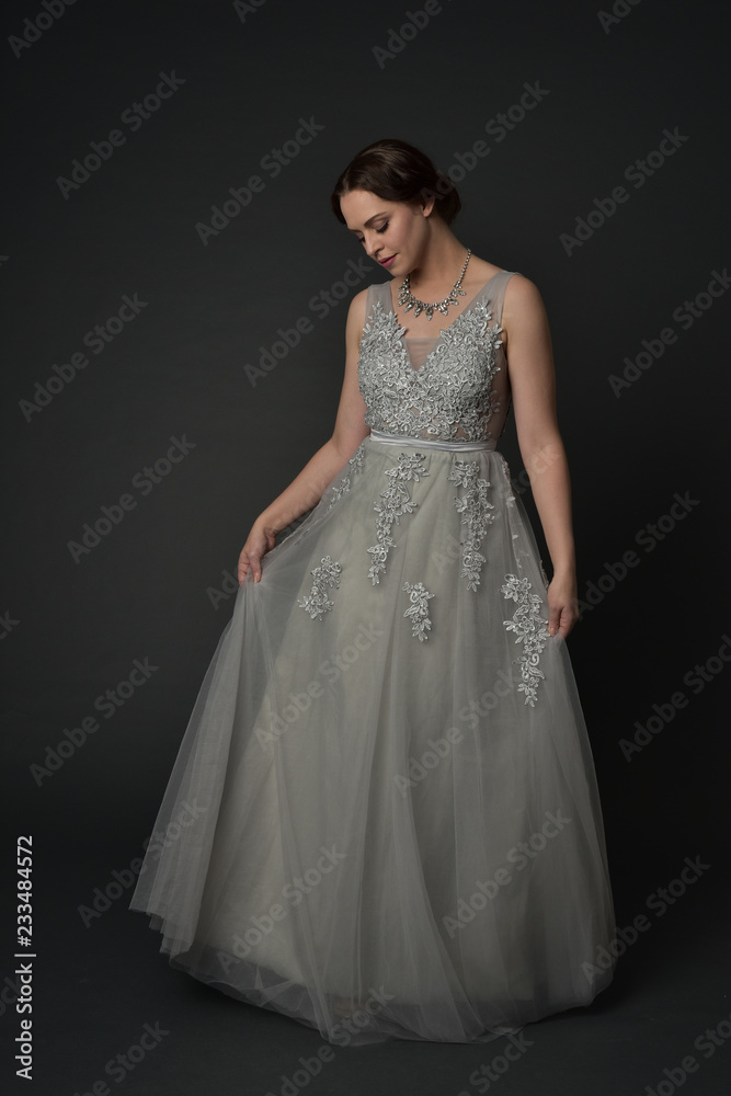 full length portrait of brunette  girl wearing long silver ball gown. standing pose on grey studio background.