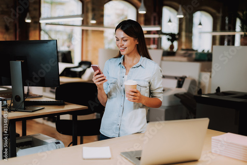 Waist up portrait of charming woman using smartphone while holding cup of coffee. She is standing near desk with laptop and smiling