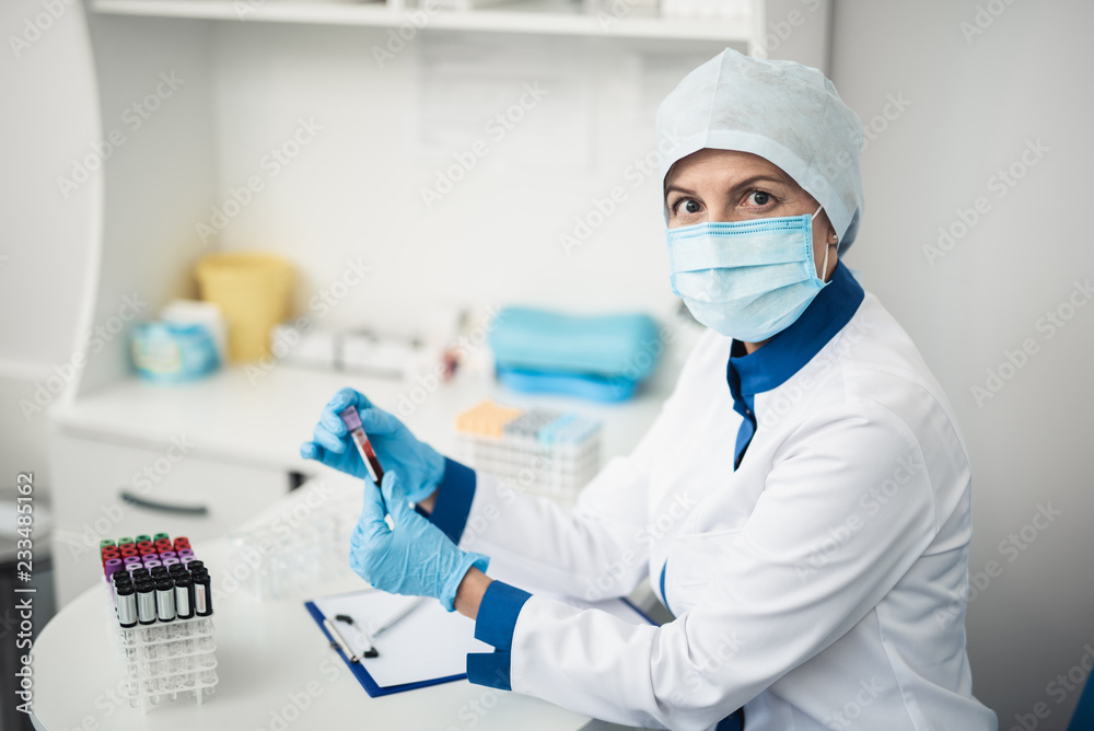 Fototapeta premium Professional laboratory research in healthcare system. Female facility assistant looking at camera while holding test tube of blood in lab office