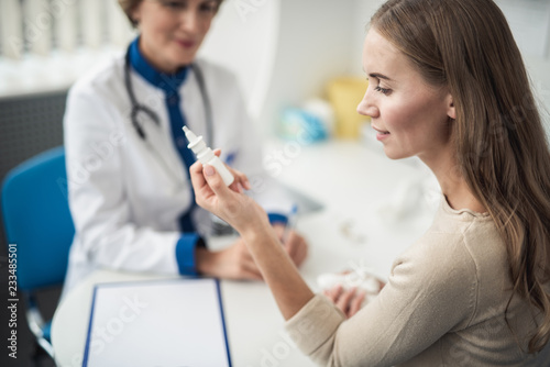 Concept of professional consultation and remedy. Waist up portrait of young female patient with running nose holding nasal drops sitting in medical office