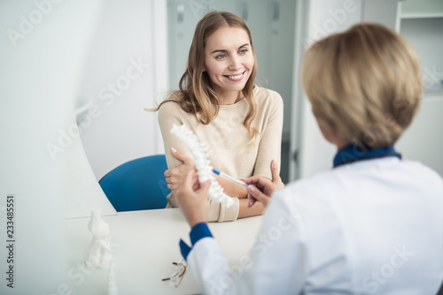 Concept of professional consultation in healthcare system. Waist up portrait of female doctor showing medical spine model to smiling young lady in clinic