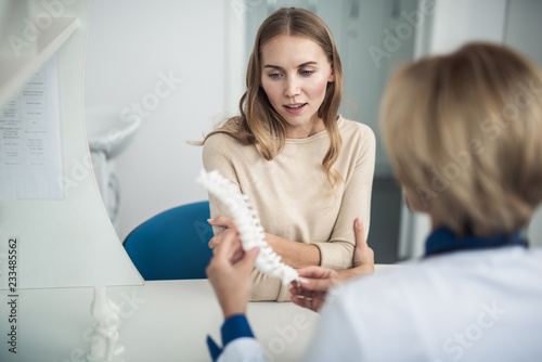 Concept of professional consultation in healthcare system. Waist up portrait of female doctor showing medical spine model to young lady in clinic
