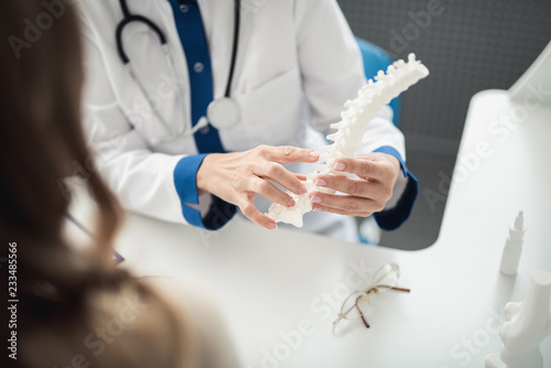 Concept of professional consultation in healthcare system. Close up portrait of female doctor showing medical spine model to young lady in clinic