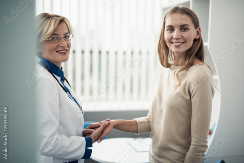 Concept of positive method of consultation in healthcare system. Waist up portrait of cheerful doctor woman holding hands of young female looking straight in camera photo