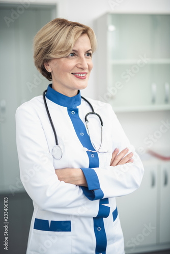 Concept of professional inspiration in healthcare system. Side on portrait of smiling female doctor in medical uniform confidently standing in cabinet crossing her arms