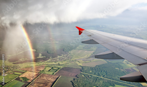 passenger airplane window seat wing view on dramatic thunderstorm rain clouds with rainbow against green rural scenery air travel aerial satellite panoramic transportation landscape background