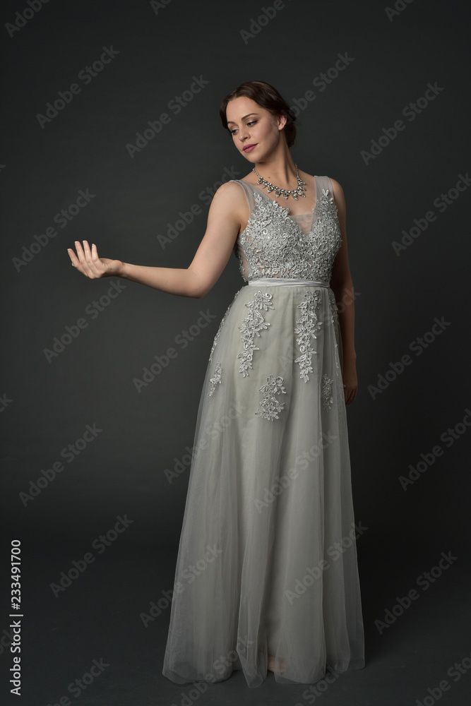 full length portrait of brunette  girl wearing long silver ball gown. standing pose on grey studio background.