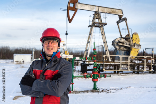 Man worker in the oilfield near pump jack and wellhead, wearing helmet and work clothes. Industrial site background. Oil and gas concept.