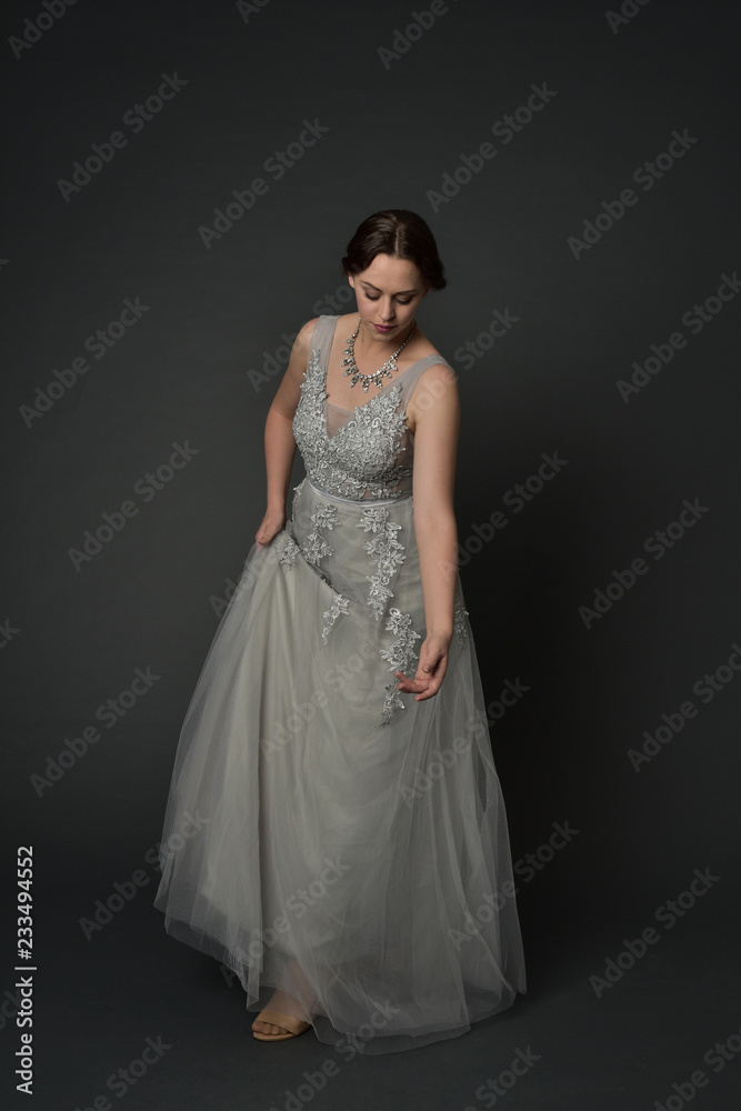 full length portrait of brunette  girl wearing long silver ball gown. standing pose on grey studio background.