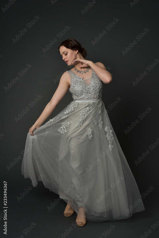 full length portrait of brunette  girl wearing long silver ball gown. standing pose on grey studio background.