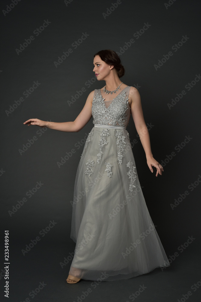 full length portrait of brunette  girl wearing long silver ball gown. standing pose on grey studio background.