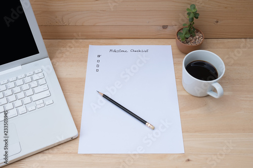 Paper check list for planning milestone, laptop, coffee on wooden table.