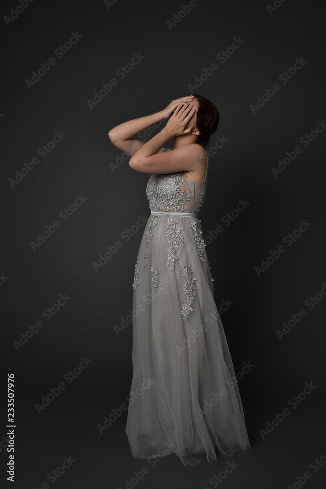 full length portrait of brunette girl wearing long silver ball gown. standing pose with hands covering face on grey studio background. full length portrait of brunette girl wearing long silver ball gown. standing pose with hands covering face on grey studio background.