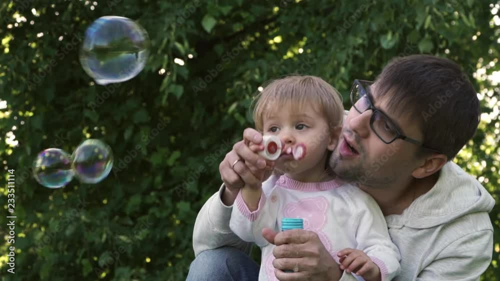 Father and little baby girl blow soap bubbles in summer park. Happy family play outdoors on weekends. Father and little baby girl blow soap bubbles in summer park. Happy family play outdoors on weekends.