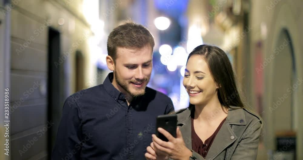 Front view of a happy couple browsing smart phone walking in the night in the street Front view of a happy couple browsing smart phone walking in the night in the street