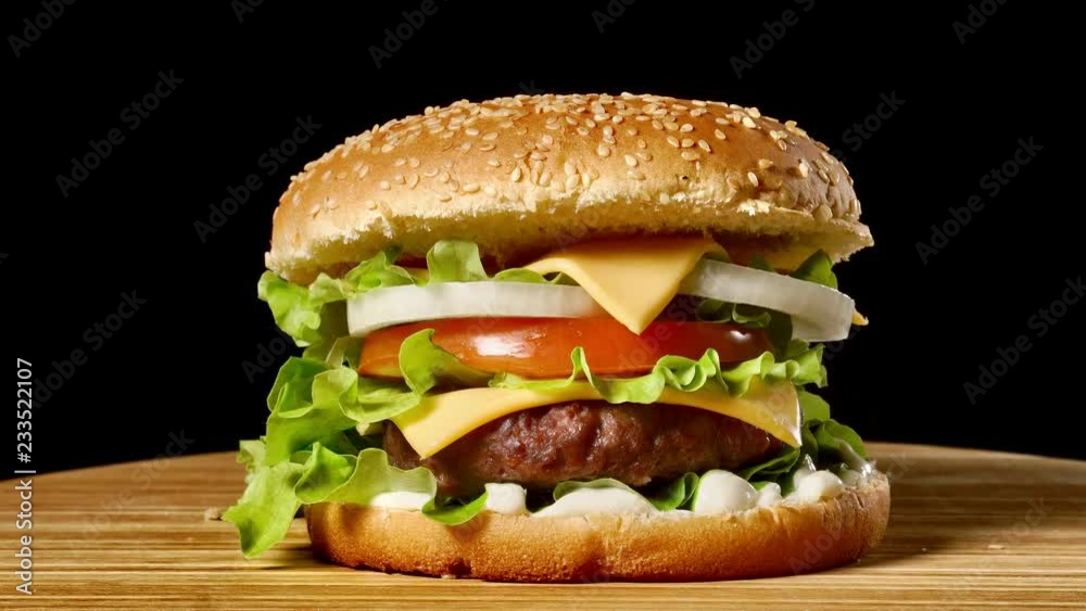 Great Burger with beef cutlet, tomatoes, mushrooms and cucumbers with melted cheese rotates on a wooden Board on black background. Great Burger with beef cutlet, tomatoes, mushrooms and cucumbers with melted cheese rotates on a wooden Board on black background.