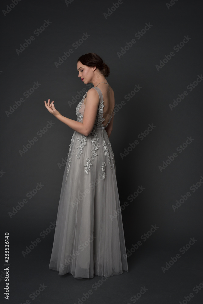 full length portrait of brunette  girl wearing long silver ball gown. standing pose on grey studio background.