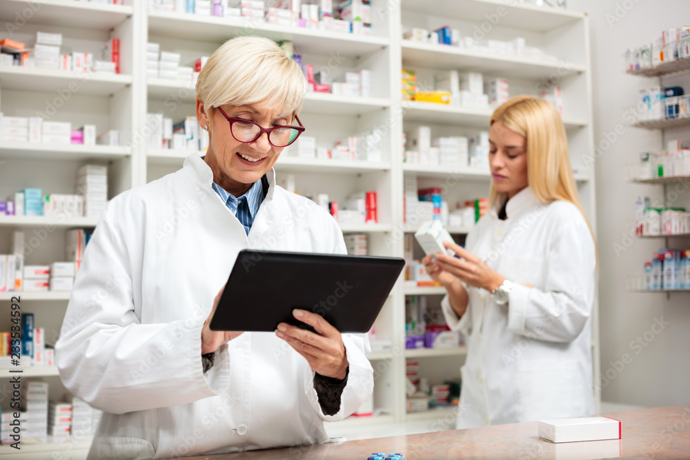 Mature and young female pharmacist working together. Mature woman is using a tablet while young woman reads labels on medication boxes. Medicine, pharmaceutics, health care and people concept