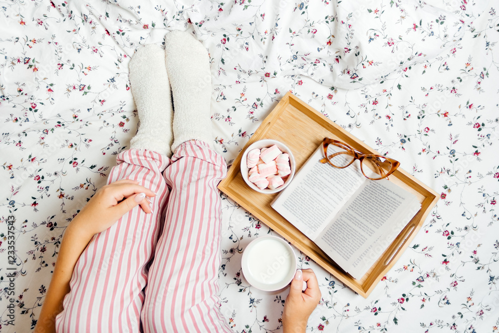 Young woman in pajamas and socks drinking milk at home in bed and reading book, top view - obrazy, fototapety, plakaty 