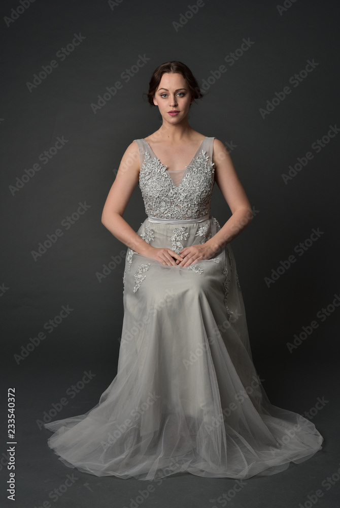  full length portrait of brunette  girl wearing long silver ball gown. seated pose on grey studio background.