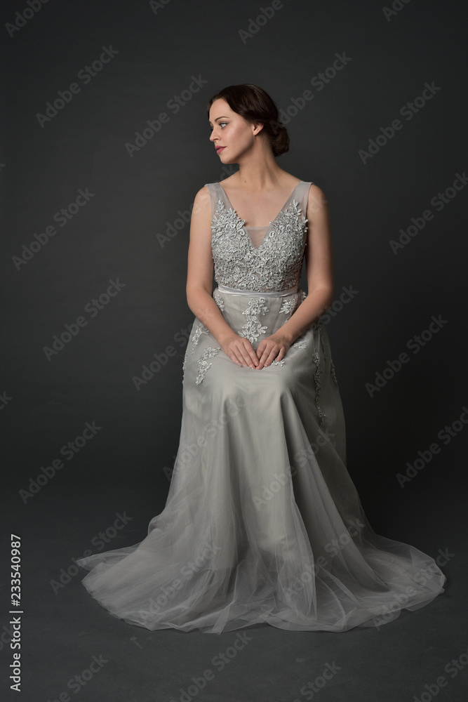 full length portrait of brunette girl wearing long silver ball gown. seated pose on grey studio background. full length portrait of brunette girl wearing long silver ball gown. seated pose on grey studio background.