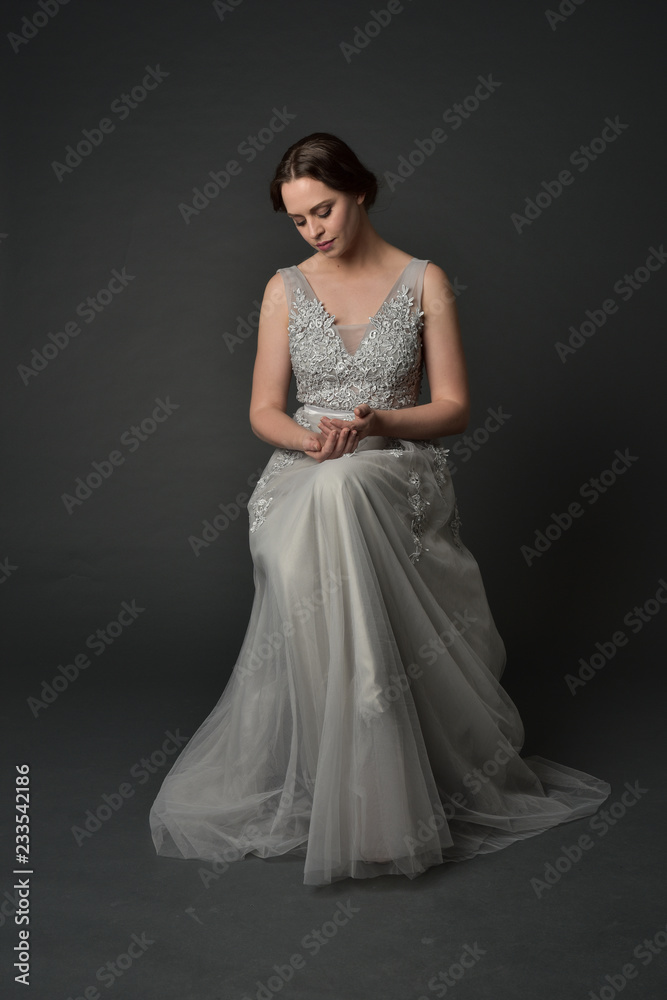 full length portrait of brunette girl wearing long silver ball gown. seated pose on grey studio background. full length portrait of brunette girl wearing long silver ball gown. seated pose on grey studio background.