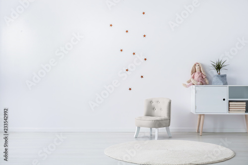 Pastel grey armchair next to wooden cabinet with books, toy and green plant in grey pot, copy space and golder stars stickers on empty white wall photo