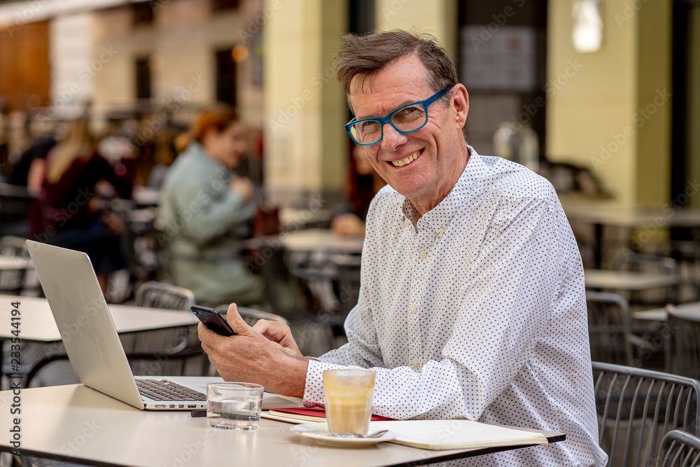 Fototapeta premium Happy attractive stylish mature man sending message on mobile while working on laptop outside cafe