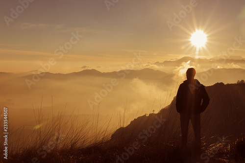 Man on top of mountain. Hiker climbed on peak of rock above foggy valley. Man watch over misty and foggy morning valley to bright morning Sun.