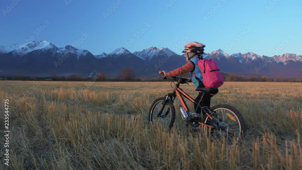 One caucasian children walk with bike in wheat field. Girl walking black orange cycle on background of beautiful snowy mountains. Biker motion ride with backpack and helmet. Mountain bike hardtail.