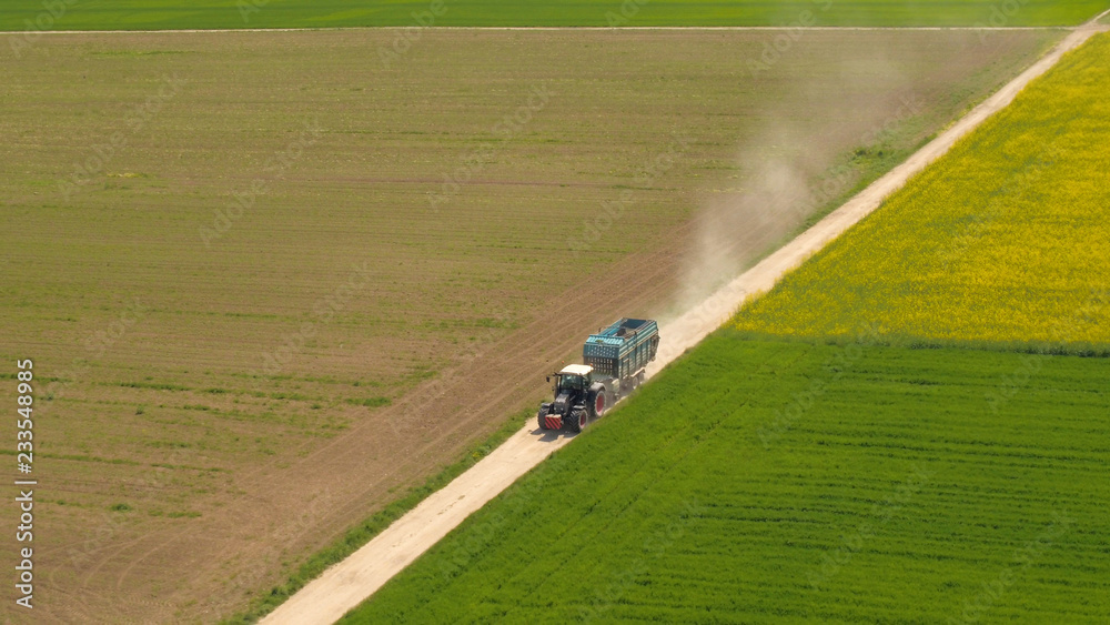 AERIAL: Tractor driving on dusty country road between colorful crop fields AERIAL: Tractor driving on dusty country road between colorful crop fields