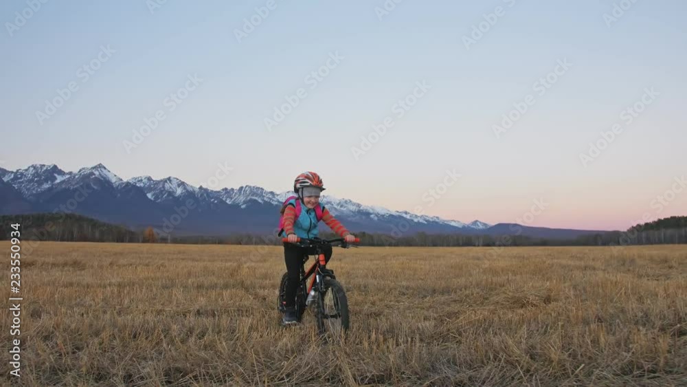 One caucasian children rides bike in wheat field. Little girl riding black orange cycle on background of beautiful snowy mountains. Biker motion ride with backpack and helmet. Mountain bike hardtail. One caucasian children rides bike in wheat field. Little girl riding black orange cycle on background of beautiful snowy mountains. Biker motion ride with backpack and helmet. Mountain bike hardtail.