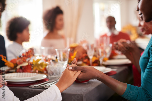 Young black adult woman and her daughter holding hands and saying grace with their multi generation family at the Thanksgiving dinner table, detail, focus on foreground photo