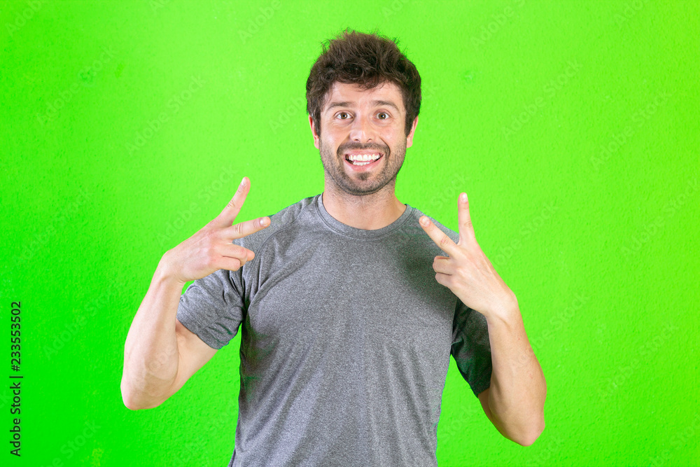 Young handsome man over green background smiling looking to the camera showing fingers doing victory sign. Number two..