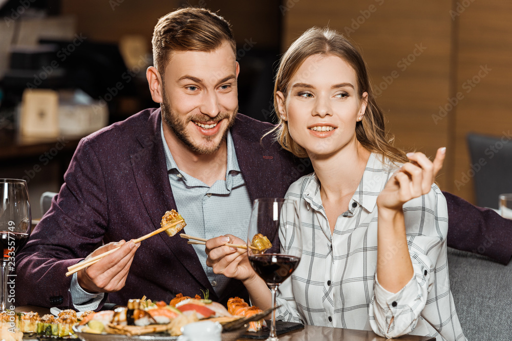 Smiling young adult couple eating sushi rolls in restaurant