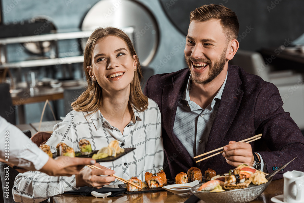 Smiling couple eating sushi rolls while waiter bringing new order in restaurant