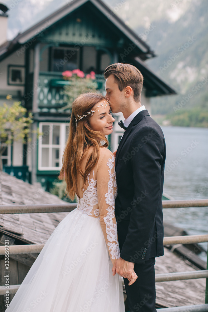 Naklejka premium A beautiful wedding couple walks on the lake and mountains background in a fairy Austrian town, Hallstatt.