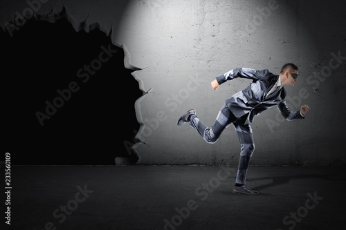 A fit young businessman in a striped suit and a black mask running away from a black hole in a wall.