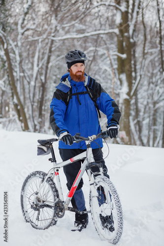 Determined young man standing next to a mountain bike, preparing for a ride in snow covered forest. Active sport lifestyle in cold weather.