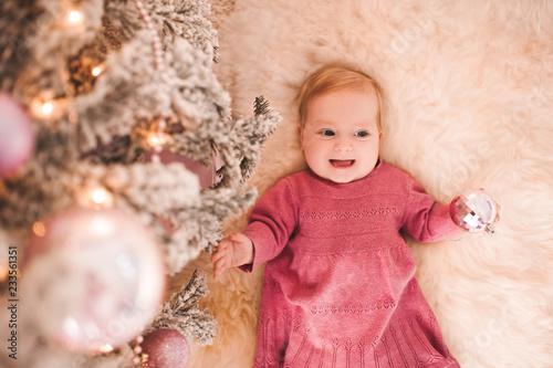 Laughing baby girl lying under Christmas tree holding Christmas ball closeup. Winter holidays. Childhood.