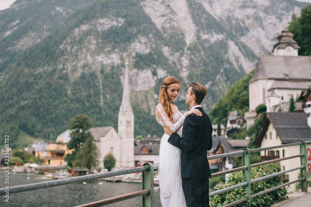 Naklejka premium A beautiful wedding couple walks on a background old cathedral, lake and mountains in a fairy Austrian town, Hallstatt.