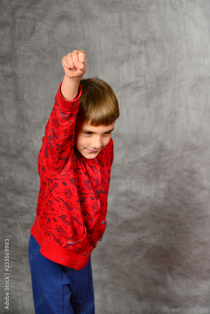 A boy in a red sweater and blue pants is standing with a raised hand up and a clenched fist in the studio on a gray background. A boy in a red sweater and blue pants is standing with a raised hand up and a clenched fist in the studio on a gray background.