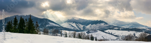 panorama of a mountainous countryside in winter. small forest on the hill and village down in the valley. snowy tops of the ridge beneath the gorgeous cloudy sky