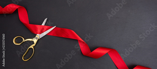 Scissors cutting red silk ribbon against black background, banner. photo