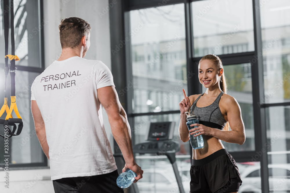 Obraz premium trainer and sportswoman holding sport bottles with water and looking at each other in gym