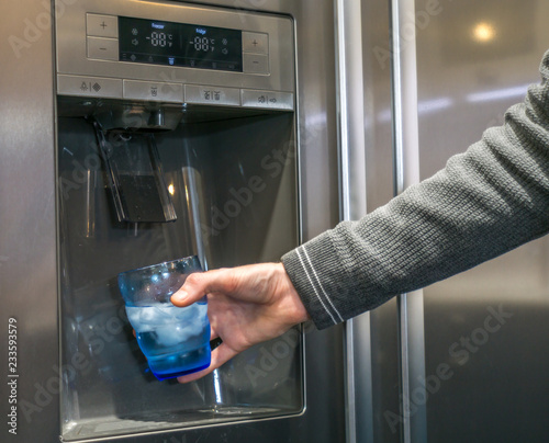 Male hand is pouring cold water and ice cubes from dispenser of home fridge.