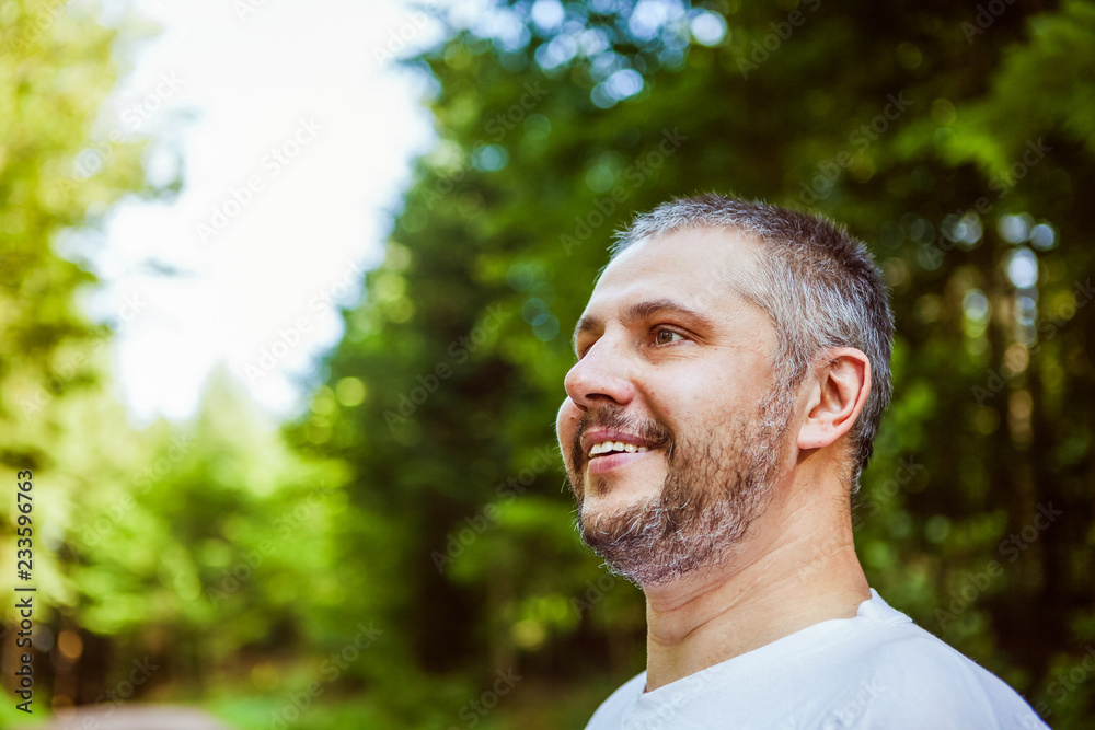 Portrait of a smiling surprised man with beard standing and looking ahead against a green trees in sunny forest Portrait of a smiling surprised man with beard standing and looking ahead against a green trees in sunny forest