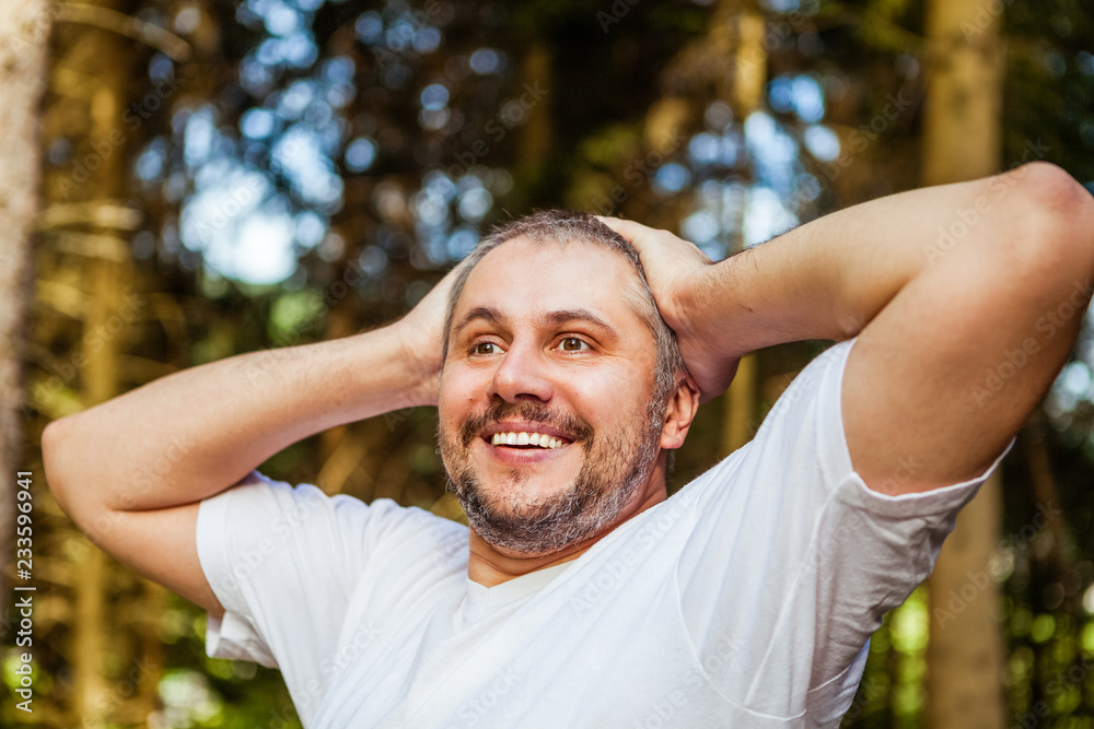 Portrait of a smiling surprised man with beard with hands up on the head looking ahead against a green trees in sunny forest Portrait of a smiling surprised man with beard with hands up on the head looking ahead against a green trees in sunny forest