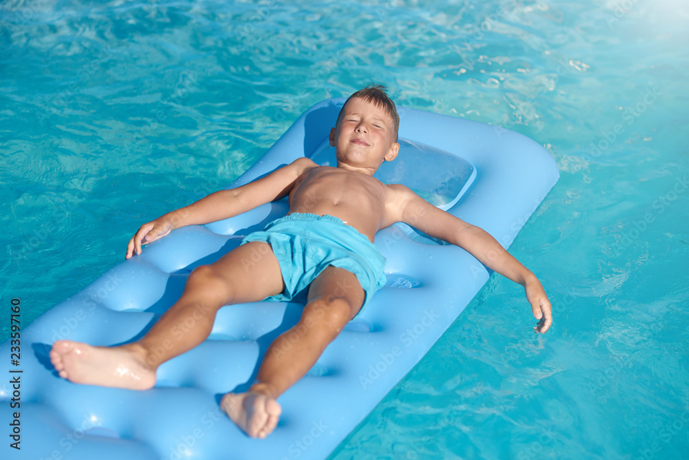 Boy relaxing at hotel swimming pool in summer. He is laying on inflatable mattress with closed eyes.